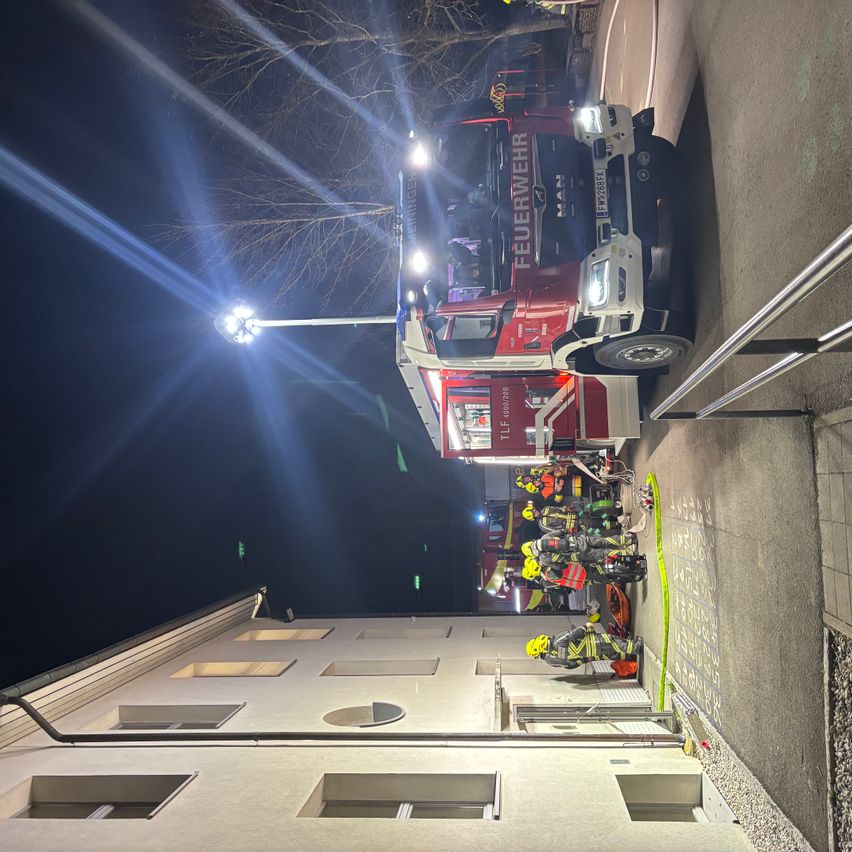 Ein roter Feuerwehrwagen mit eingeschalteten Lichtern ist an der Seite eines Gebäudes geparkt. Mehrere Feuerwehrleute arbeiten in der Nähe des Trucks an einer Aufgabe.