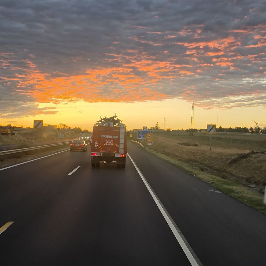 A fire truck drives on a highway with a sunset in the background, accompanied by a car. The sky is filled with clouds and the sun is setting.