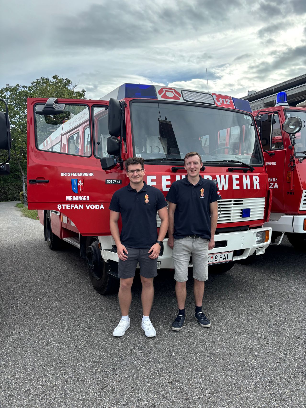 Two men stand in front of a red fire truck with the number 1324 on the side. They are both wearing black shirts and shorts. The fire truck has the word 'Feuerwehr' on it.