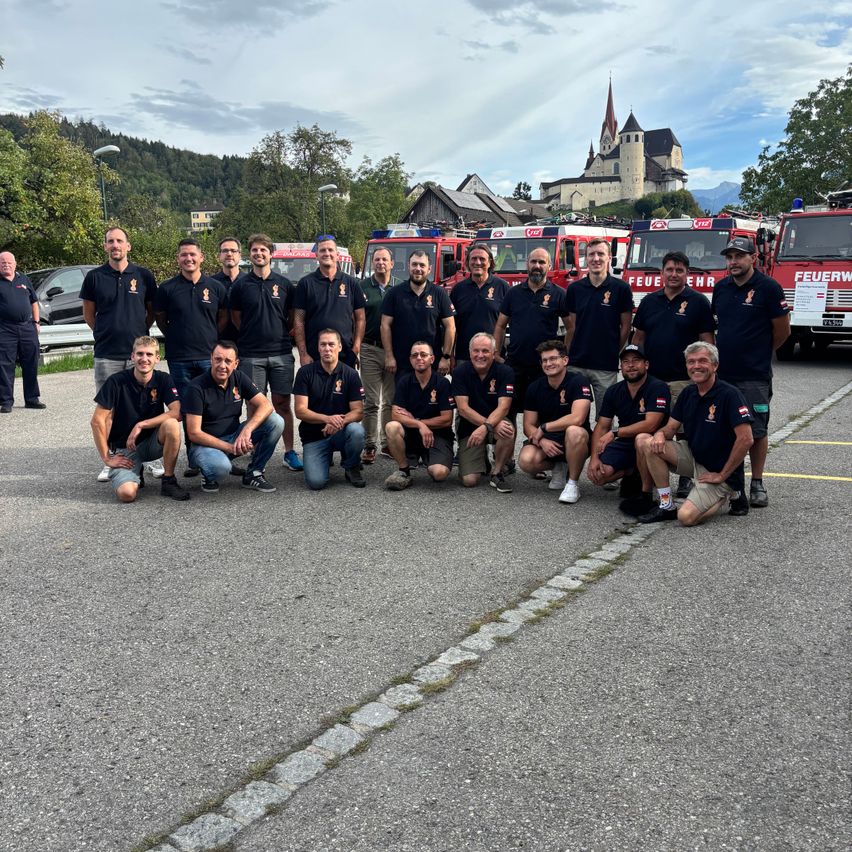 A group of firefighters in black uniforms poses for a photo in front of fire trucks.