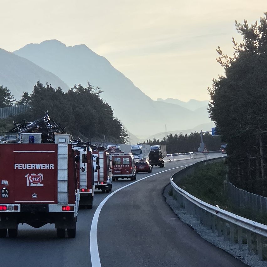 A line of red fire trucks with flashing lights are driving on a road with mountains in the background.