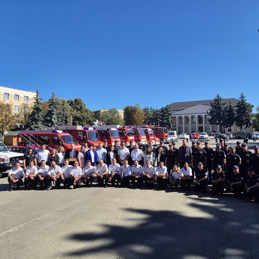Eine Gruppe von Feuerwehrleuten posiert für ein Foto vor einer Reihe von Feuerwehrwagen an einem sonnigen Tag.