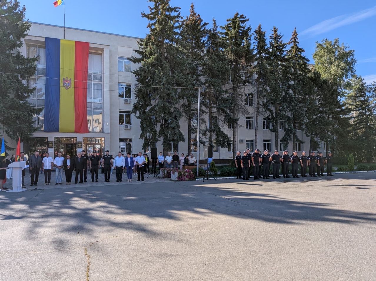 Eine Gruppe von Menschen steht vor einem Gebäude mit einer rumänischen Flagge auf der Spitze. Sie sind in einer Reihe aufgestellt und scheinen an einer Zeremonie teilzunehmen.