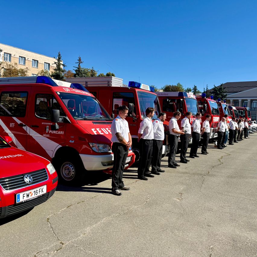 Eine Reihe roter Feuerwehrfahrzeuge mit Feuerwehrleuten davor. Die Fahrzeuge sind in einer Reihe geparkt, mit einem Gebäude im Hintergrund.