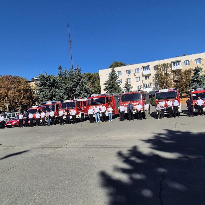 Eine Gruppe von Feuerwehrleuten in Uniform steht vor ihren Feuerwehrwagen auf einem Parkplatz.