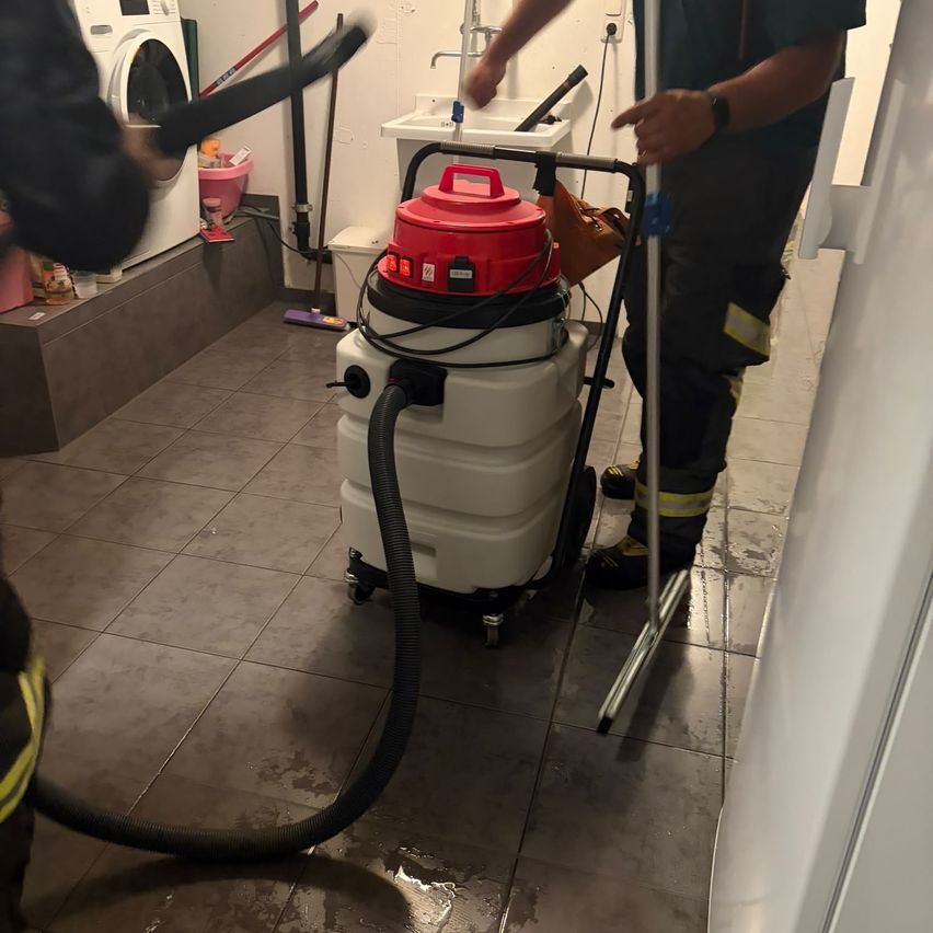 Zwei Personen benutzen einen rot-weißen Staubsauger, um einen gefliesten Boden zu reinigen. Im Hintergrund ist eine Waschmaschine zu sehen.