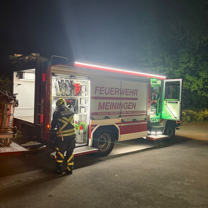 Ein Feuerwehrmann steht neben einem offenen Feuerwehrwagen mit roten Lichtern, der nachts auf einer Straße geparkt ist.