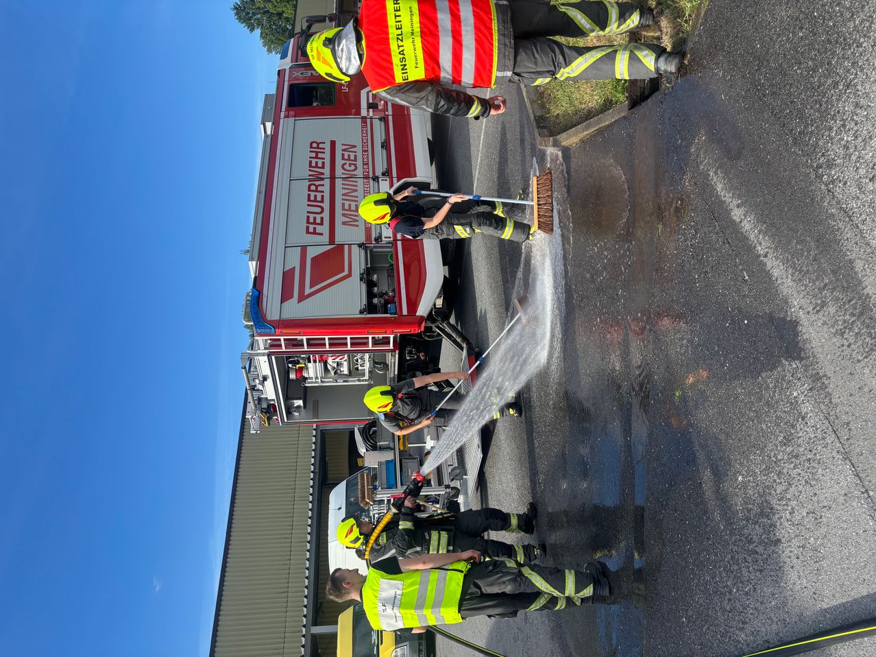 Firefighters are cleaning the street with a fire engine in the background under a clear sky.