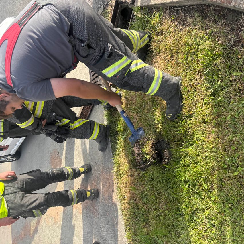 A firefighter is working to fix a drain cover on the road, while another firefighter stands nearby.