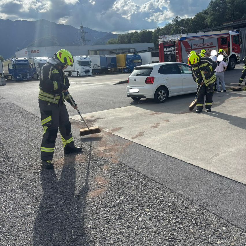 Firefighters are cleaning a parking lot, one with a broom and another with a brush, wearing yellow helmets and safety suits. A white car is parked nearby. In the background, a fire truck is parked.