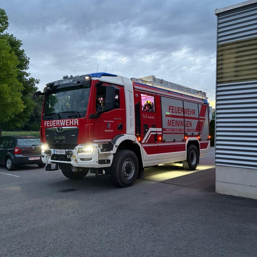 Ein roter Feuerwehrwagen mit der Aufschrift Feuerwehr Meiningen steht mit eingeschalteten Scheinwerfern auf einem Parkplatz.