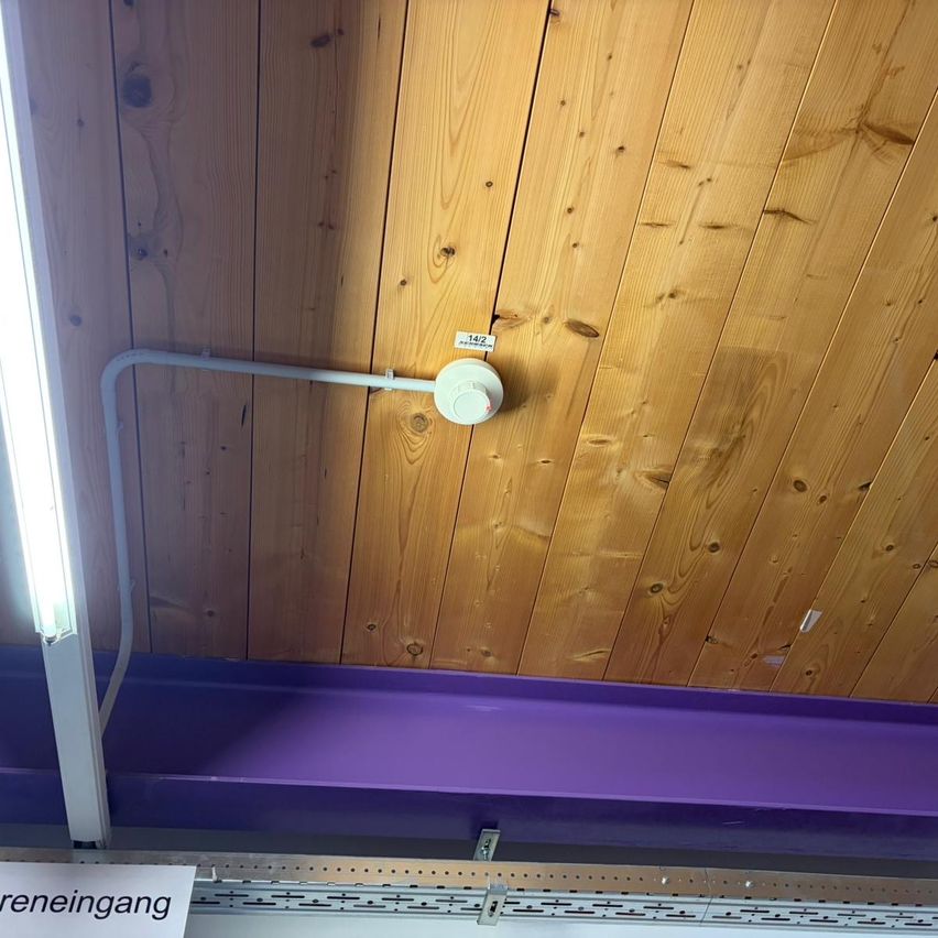 Eine Holzdecke mit einem weißen Rohr und Lichtanlage befestigt. Das Rohr hat einen weißen Deckel und ein Etikett. Darunter befindet sich ein violettes Regal mit einem weißen Schild, das 'Reinigung' besagt.