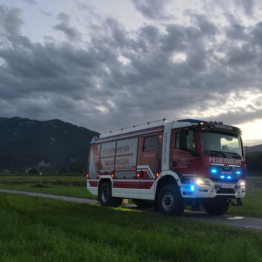 Ein Feuerwehrwagen aus Meiningen fährt auf einer Landstraße unter einem bewölkten Himmel, mit Bergen im Hintergrund.