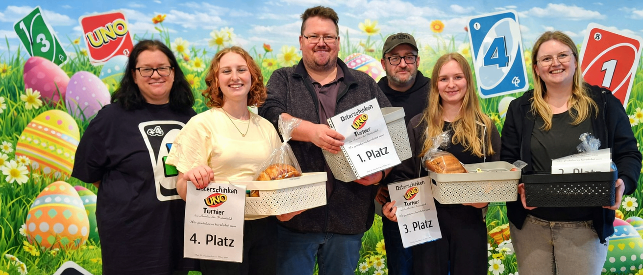 Fünf Personen stehen auf einem grünen Rasen und halten Körbe mit Essen. Sie haben nummerierte Schilder für den 1., 2., 3. und 4. Platz. Dahinter ist eine Kulisse mit einem Spielbrett, das eine Nummer 4 zeigt.