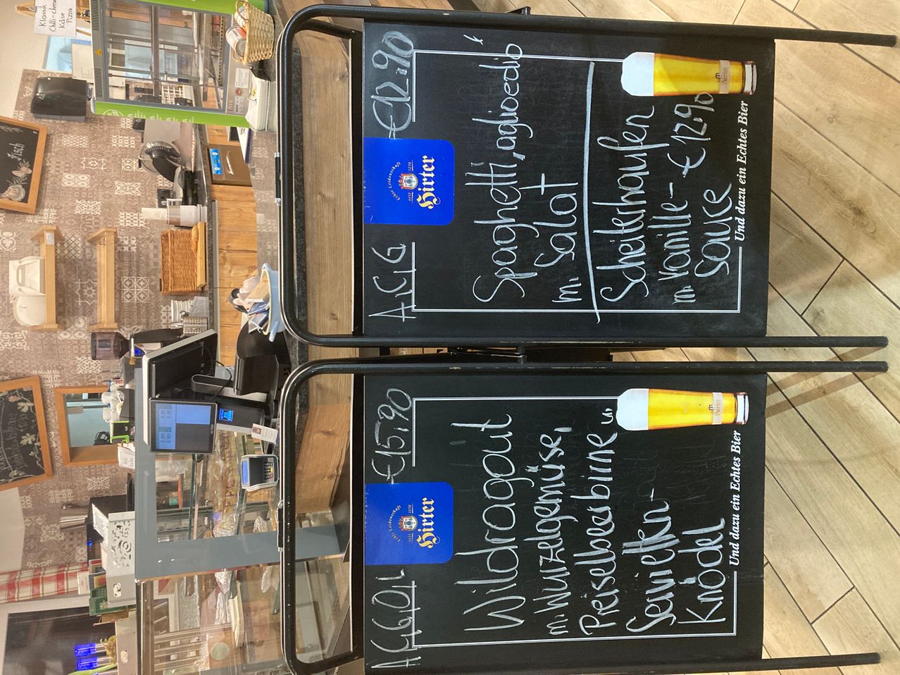 Zwei Tafeln zeigen Preise für Bier und Essen. Oben steht ein Schild 'ACG €2,90'. Darunter eine blaue Karte mit 'Sitter'. Unten auf der Tafel steht ein Schild 'ACG €3,90'. Eine blaue Karte mit 'Sitter'. Beide Tafeln haben Essensposten und Preise in Deutsch aufgeführt.