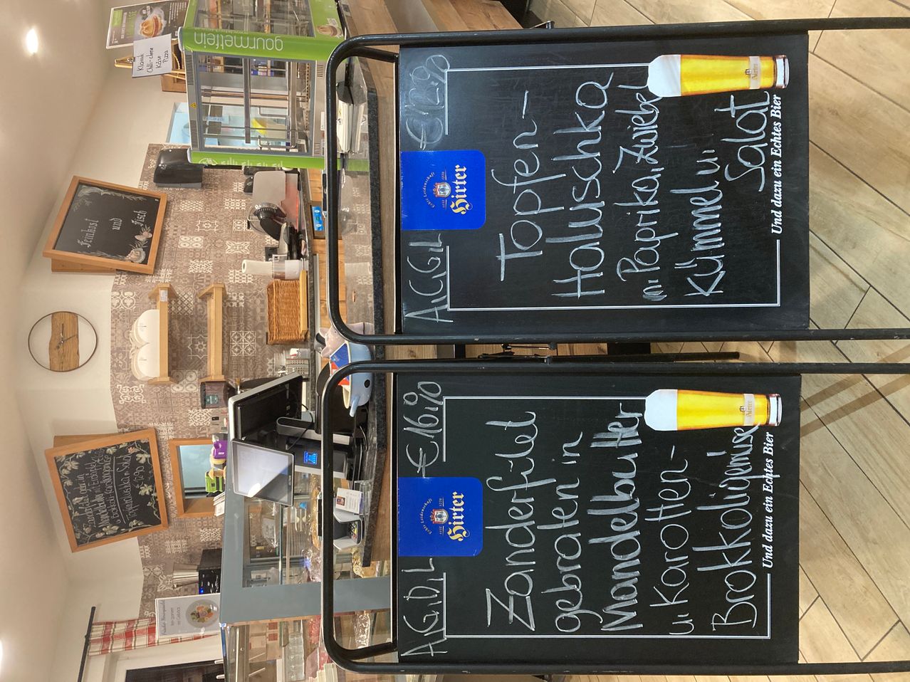 Zwei schwarze Tafeln mit darauf geschriebenen Speisekarten stehen auf einem schwarzen Wagen. Sie haben Biernamen und ein Bierglas oben drauf. Hinter den Tafeln sind Displays, ein Regal und Monitore. Es gibt einen gefliesten Boden mit einer Box und einer weißen Schüssel.