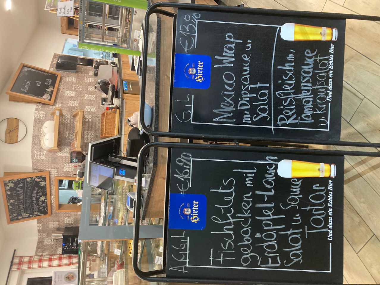 Two chalkboard menus are displayed on a metal stand. One menu lists items like 'Mexico Wrap' and 'Spicy Dip Sauce'. The other lists 'Fishcakes' and 'Mushroom Soup'. Both have blue stickers reading 'Sirtet'.