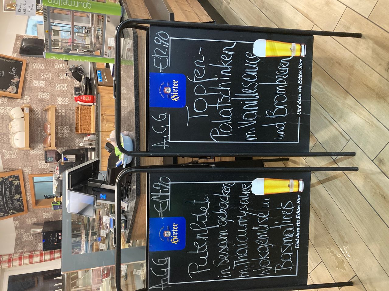 Two chalkboards display beer options, including Pilsner Urquell and Radler, with prices and descriptions in German. The background shows a shop interior.