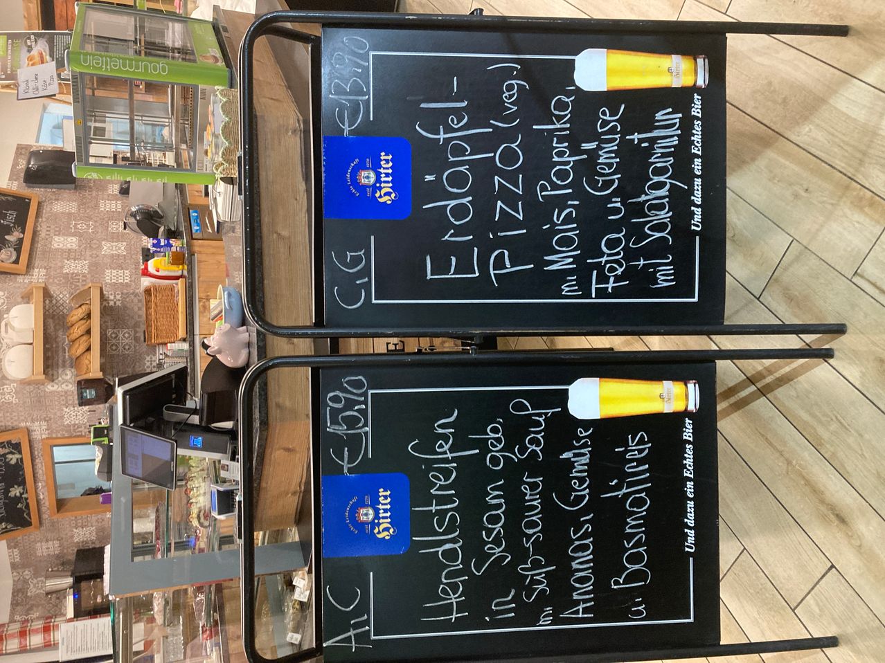 Zwei Tafeln zeigen tägliche Essens- und Getränkeangebote. Eine listet 'Etoffel-Pizza' auf, die andere 'Heidelbeerjoghurt'. Beide haben Preise und ein Bierlogo.