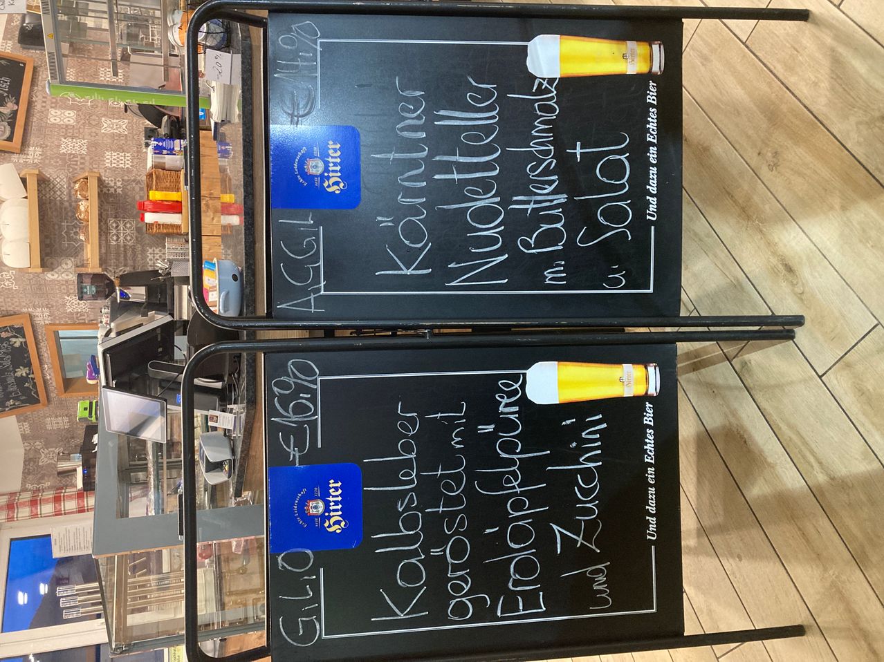 Two chalkboards with handwritten text and beer glass illustrations. The top one reads 'Kölsch' and 'Nussteller' with a price tag. The bottom one reads 'Kölschbier' and 'Erfrischungsgetränk' with a price tag.