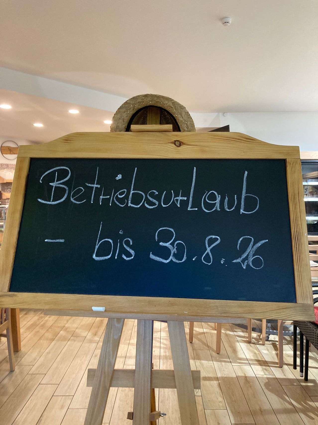 A blackboard with white chalk writing on it, displaying the message 'Betriebsurlaub - bis 30.8.26'. The blackboard is placed on a wooden easel in a room with wooden floors and chairs.