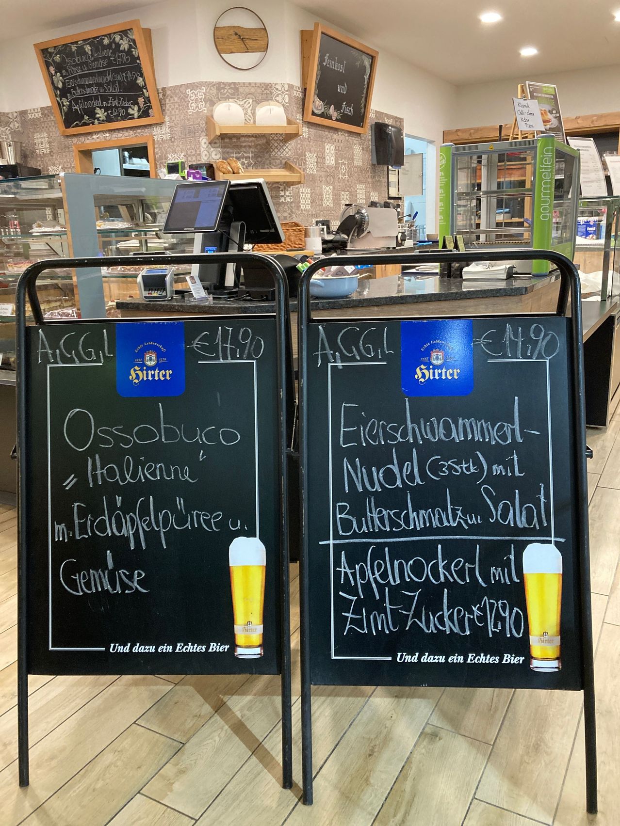Zwei Kreidetafeln mit handgeschriebenen Menüs in einem Restaurant. Die linke Tafel listet 'Ossobuco alla Milanese' mit einem Preis von 14,90 € und einer Bierempfehlung auf. Die rechte Tafel zeigt 'Eierschwammerl Nudel' mit einem Preis von 14,90 € und einer Bierempfehlung. Beide Tafeln haben ein Bierlogo und eine Bierglasillustration.