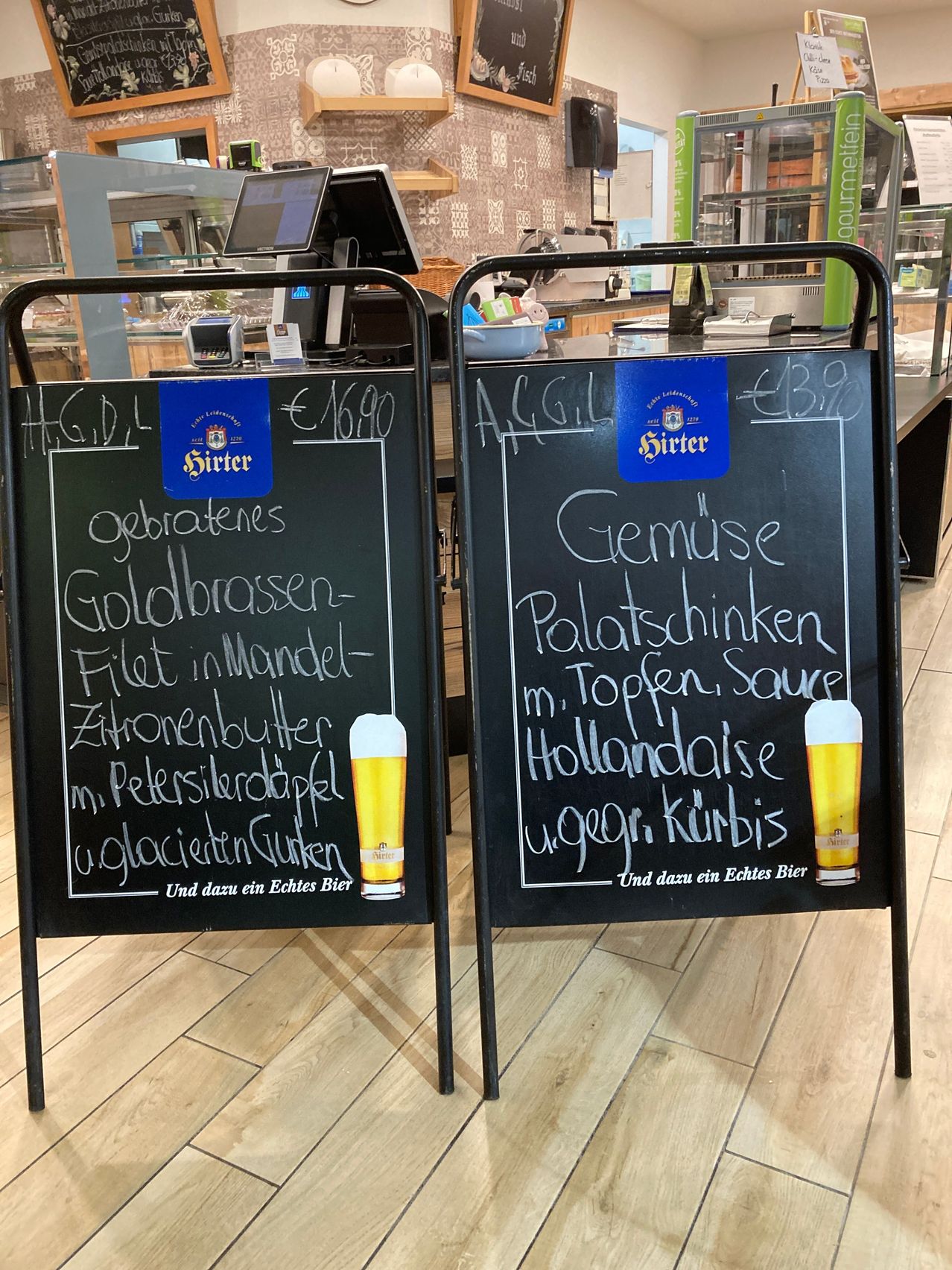 Zwei Kreidetafeln mit handgeschriebenen Menüs in einem deutschen Restaurant. Eine Tafel listet 'Gebratenes Goldbrasse-Filet in Mandel-Zitronenbutter m. Petersierdappel u.glacierten Gurken' und die andere 'Gemuese Poltoschinken m. Topfen, Sauce Hollandaise u.gegr. Kurbis'. Beide haben ein Glas Bier und einen Preisschild.
