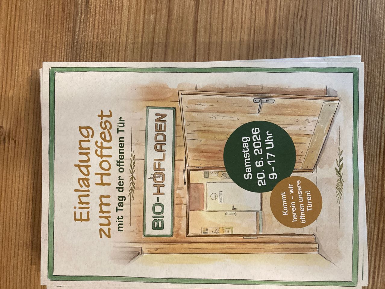 Ein Einladungstext zur Türöffnung mit einer offenen Tür, einer hölzernen Wand, einem Schloss und einem Schild, das 'Bio-Hofladden' sagt. Datum: 20.6.2026. Zeit: 9-17 Uhr.