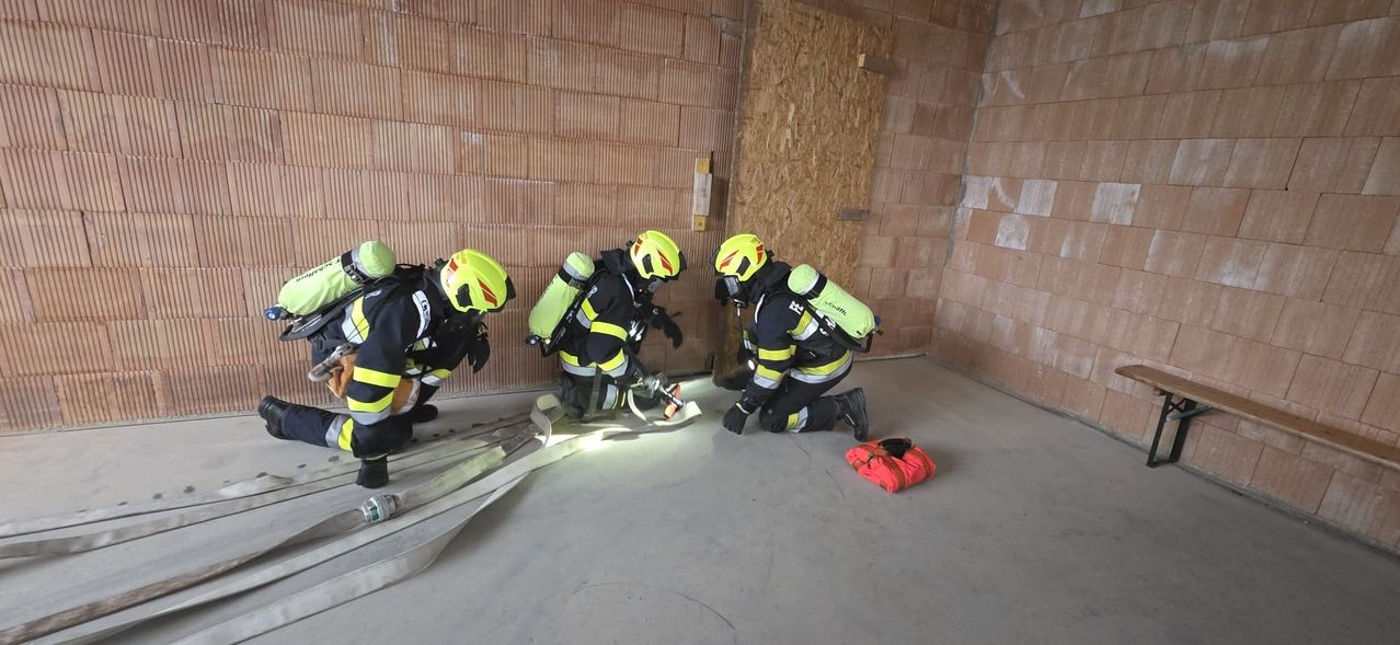 Vier Feuerwehrleute in voller Ausrüstung arbeiten in einem Schulungsraum mit Ziegelwänden, einer kniet, und die anderen stehen um eine rote Tasche herum.