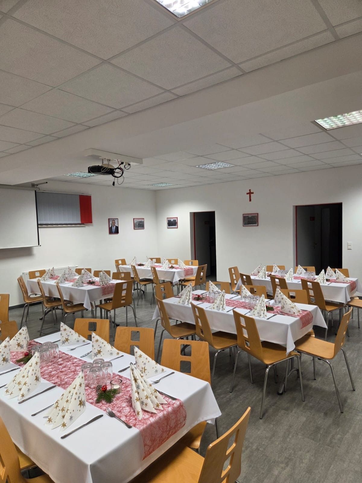 Ein leerer Bankettsaal mit Tischen und Stühlen für eine besondere Veranstaltung. Die Tische sind mit weißen Tüchern bedeckt und mit roten Tüchern und Servietten verziert. Die Stühle sind in einer Reihe angeordnet, und der Raum hat einen Projektor und eine Tafel. Es gibt auch Bilderrahmen an der Wand angebracht.