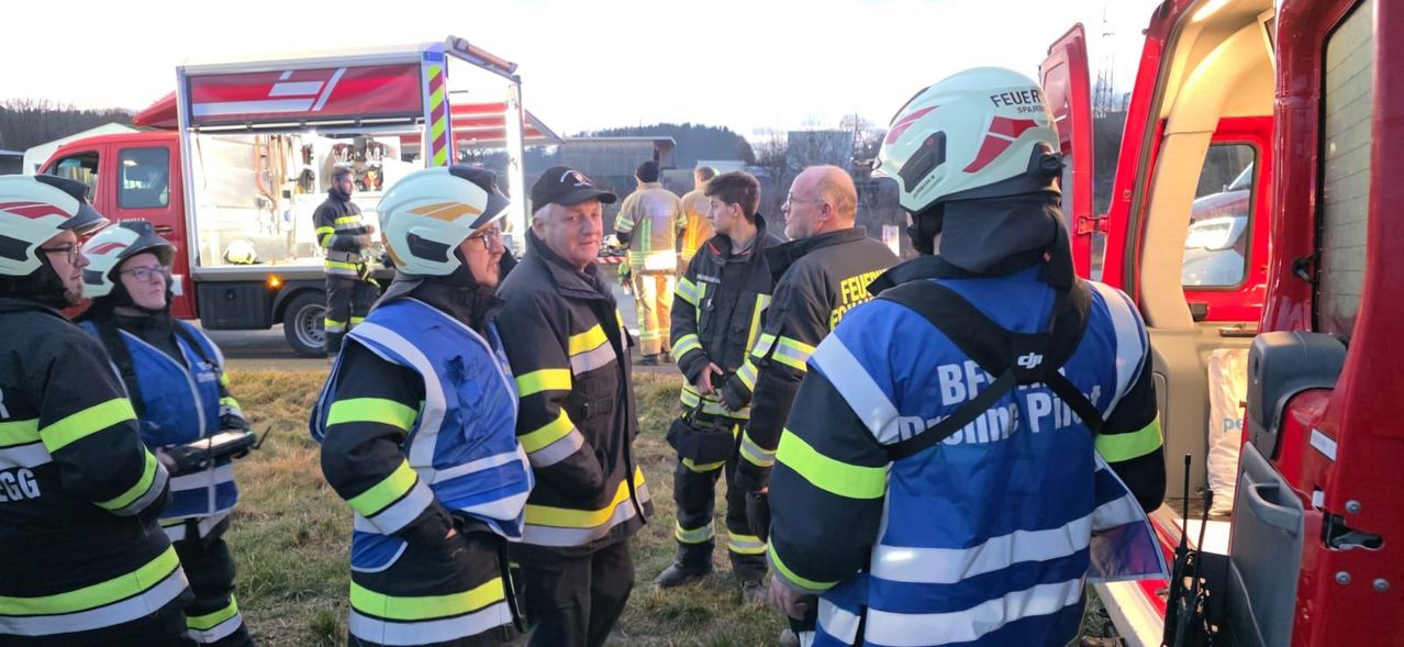 A group of firefighters stands outside, two wearing helmets, one with a cap, and another with glasses. A fire truck is behind them.