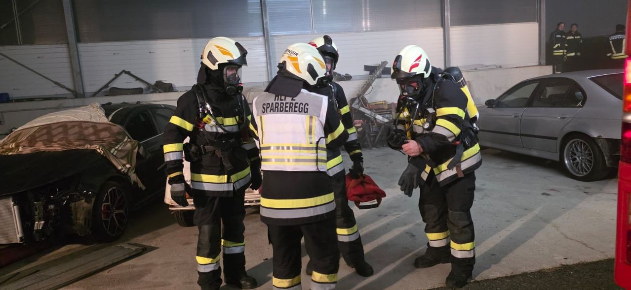 Four firefighters in full gear, including helmets and face masks, stand inside a building. One holds a red bag. The man with 'HLF' on his back wears a white vest.