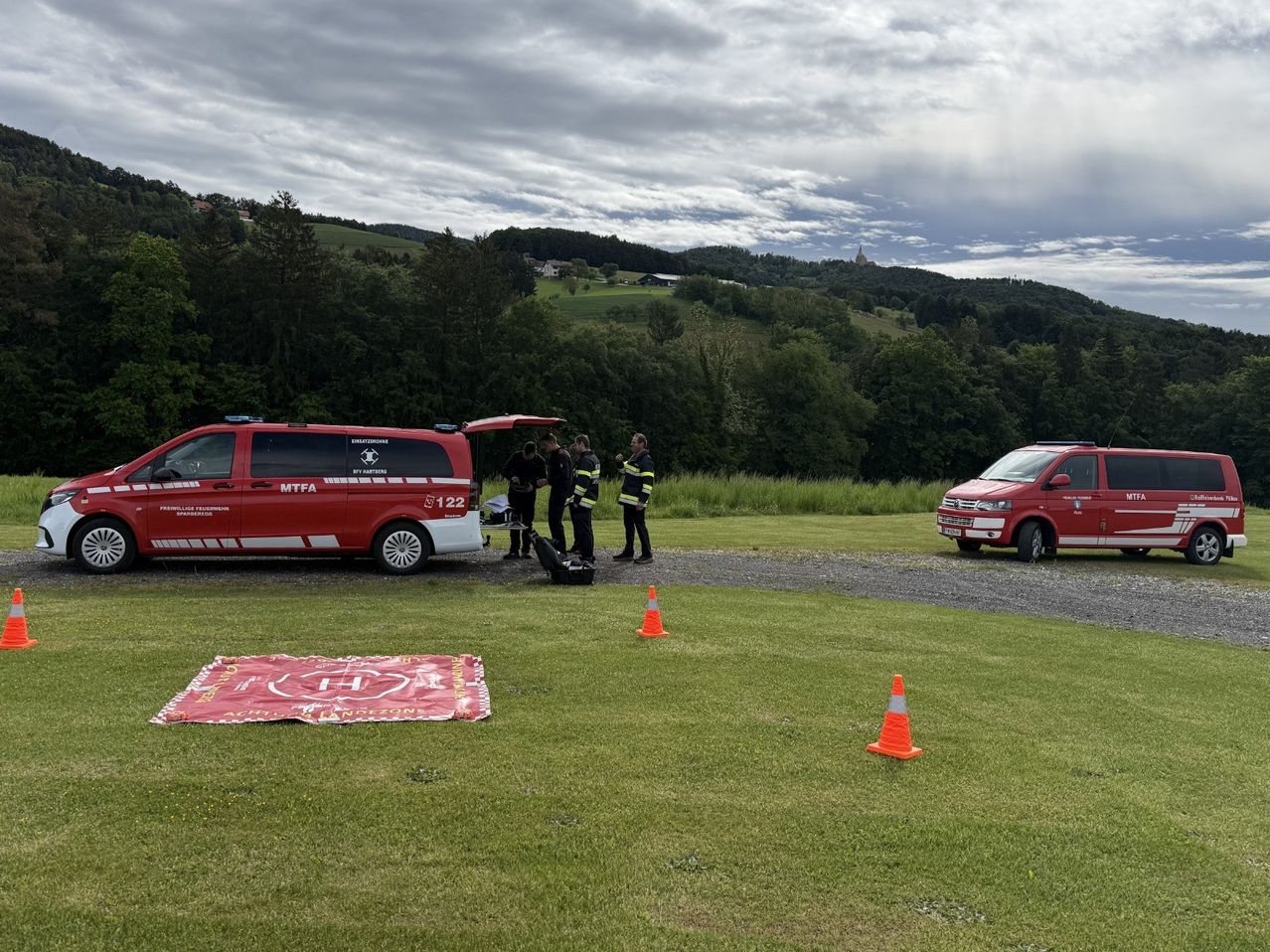 Two red vans are parked on a grassy area, one with its back door open. A group of people, possibly rescue workers, are standing around a table with equipment. A red cloth is on the ground with a logo, and orange cones are placed nearby. Trees and a mountain are in the background.