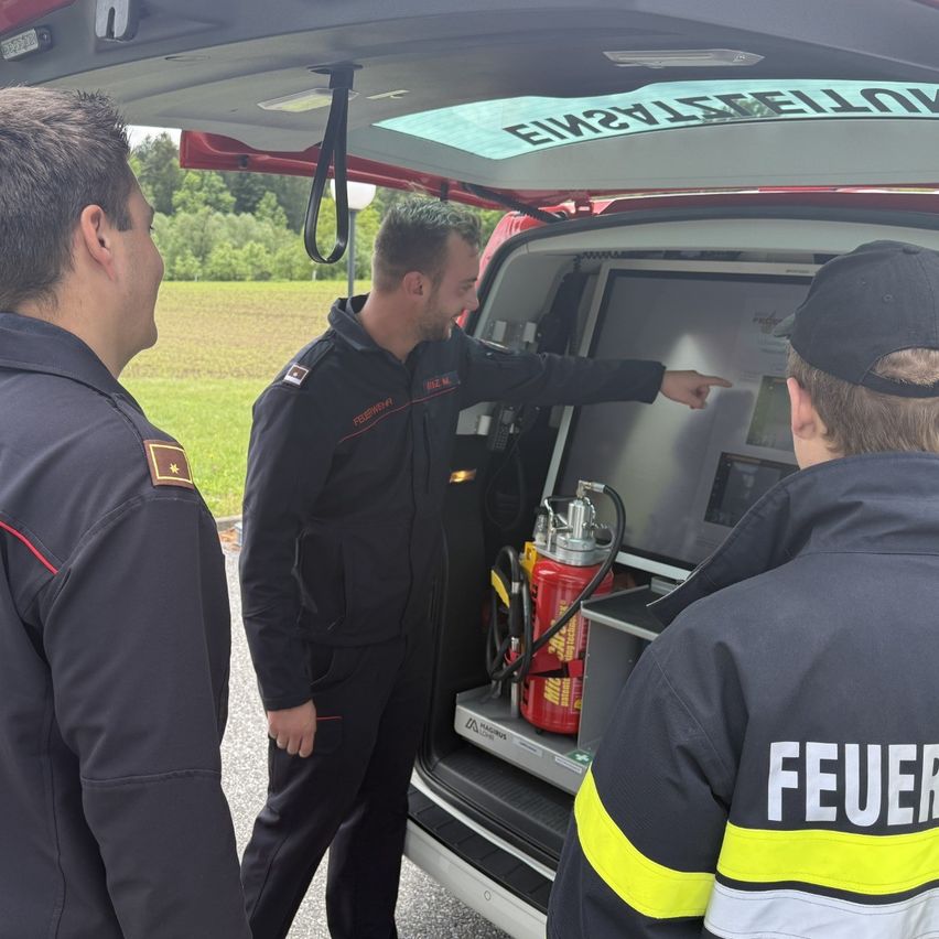 Three firefighters are standing by a vehicle with its back door open. The man in the middle is pointing at the screen, while the others look on. The vehicle has a red extinguisher on the floor.