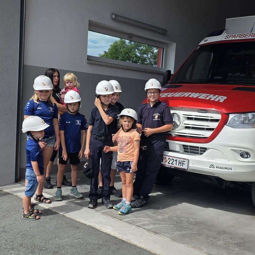Eine Gruppe von Kindern und Erwachsenen, einige mit Helmen, stehen vor einem roten und weißen Feuerwehrauto. Die Kinder posieren für ein Foto.