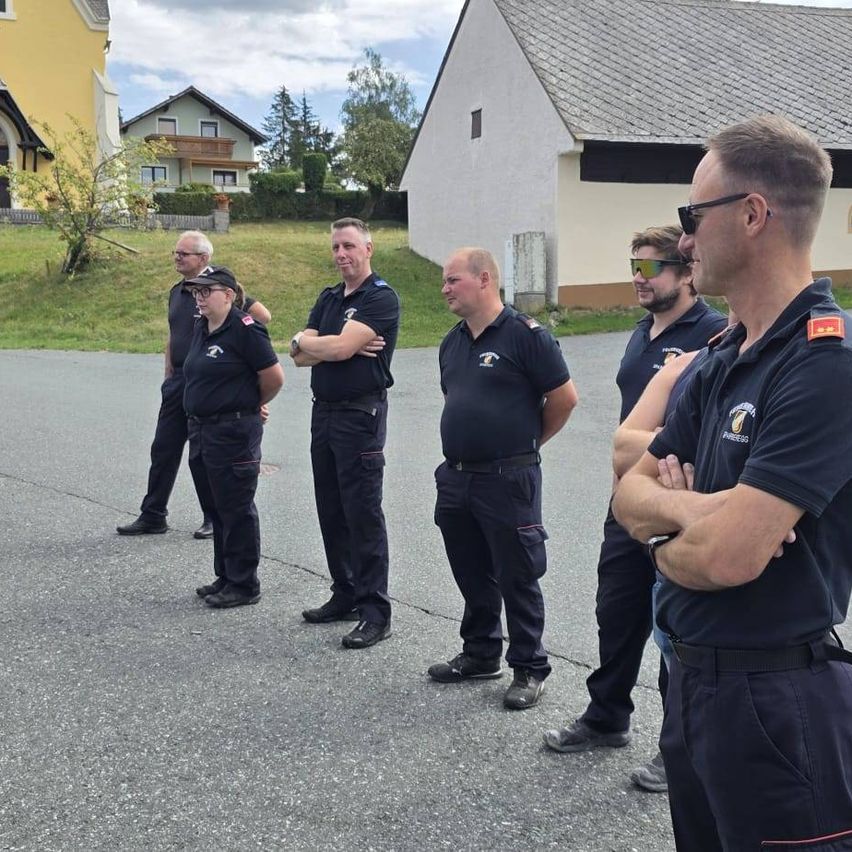 Eine Gruppe von Feuerwehrleuten in Uniform steht in einer Reihe auf einer Straße, mit einem Haus und einem Rasen im Hintergrund.