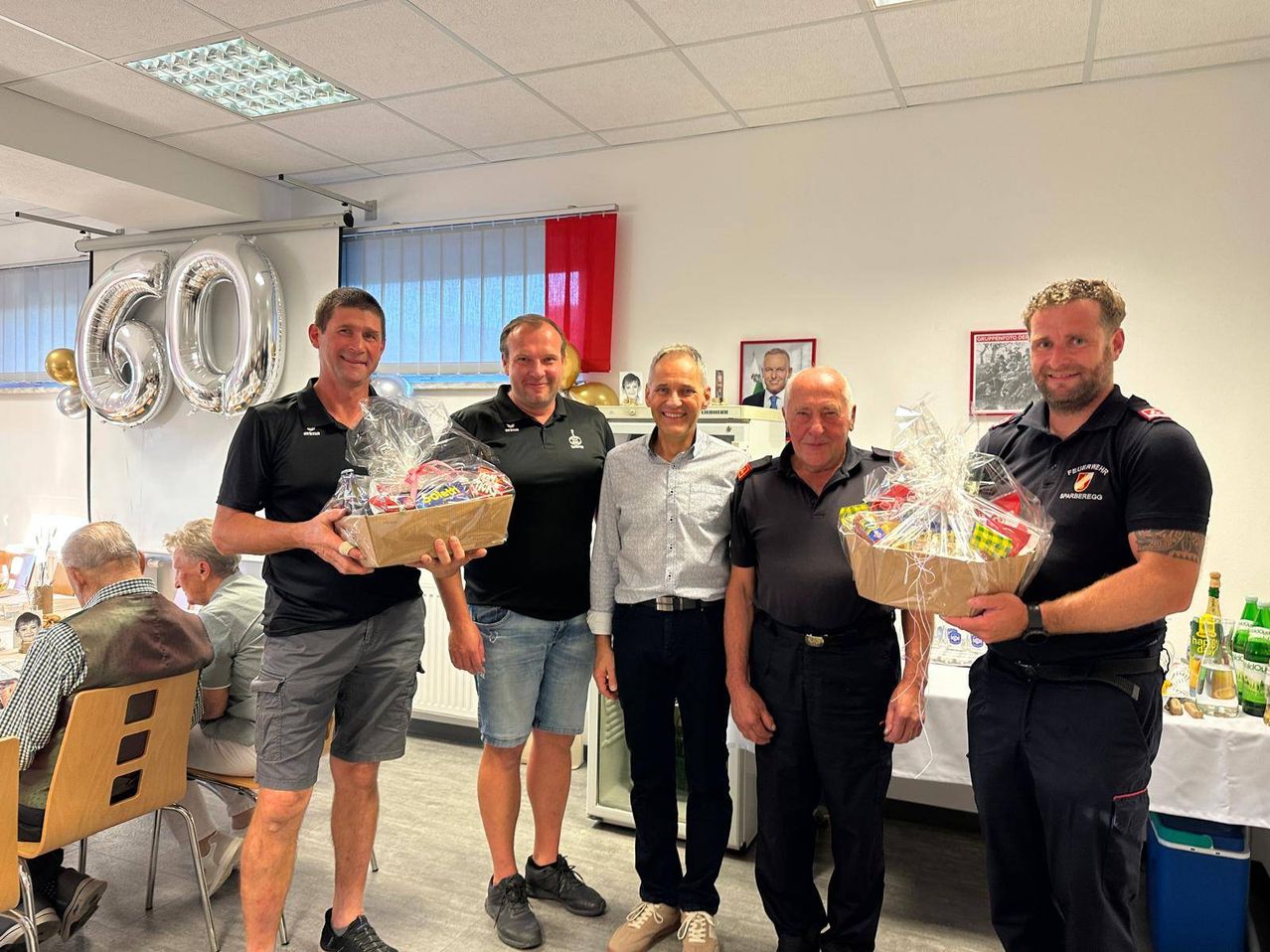 Five men stand in a room, smiling and holding gift baskets. Behind them, a wall displays a red curtain, a picture frame, and a balloon. The room is well-lit with white walls and ceiling.