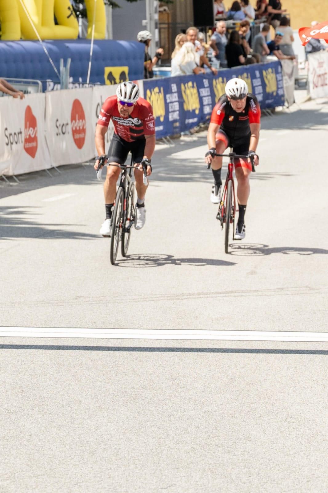 Two cyclists race down a street, both wearing helmets and cycling gear. Banners line the side of the road.