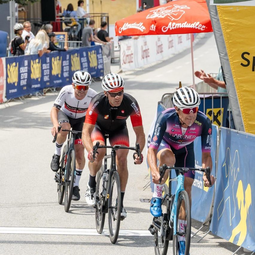 Three cyclists in cycling gear compete on a road with spectators watching from behind barriers. The background features an orange umbrella and a yellow banner.