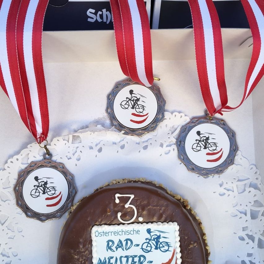 A cake in a white doily with three medals around it. The cake has a chocolate base and a white icing with text and a cyclist image.