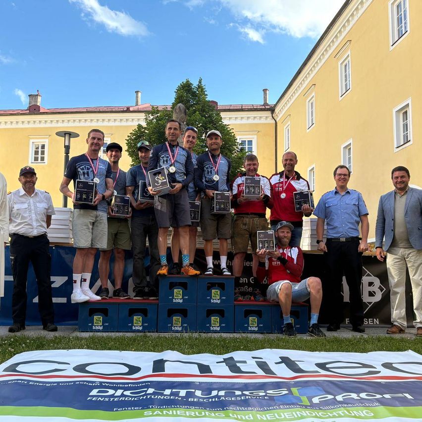 Winners of a sports event pose with medals on a stage, with a yellow building and tree in the background. A banner with the word 'contitec' lies on the ground.