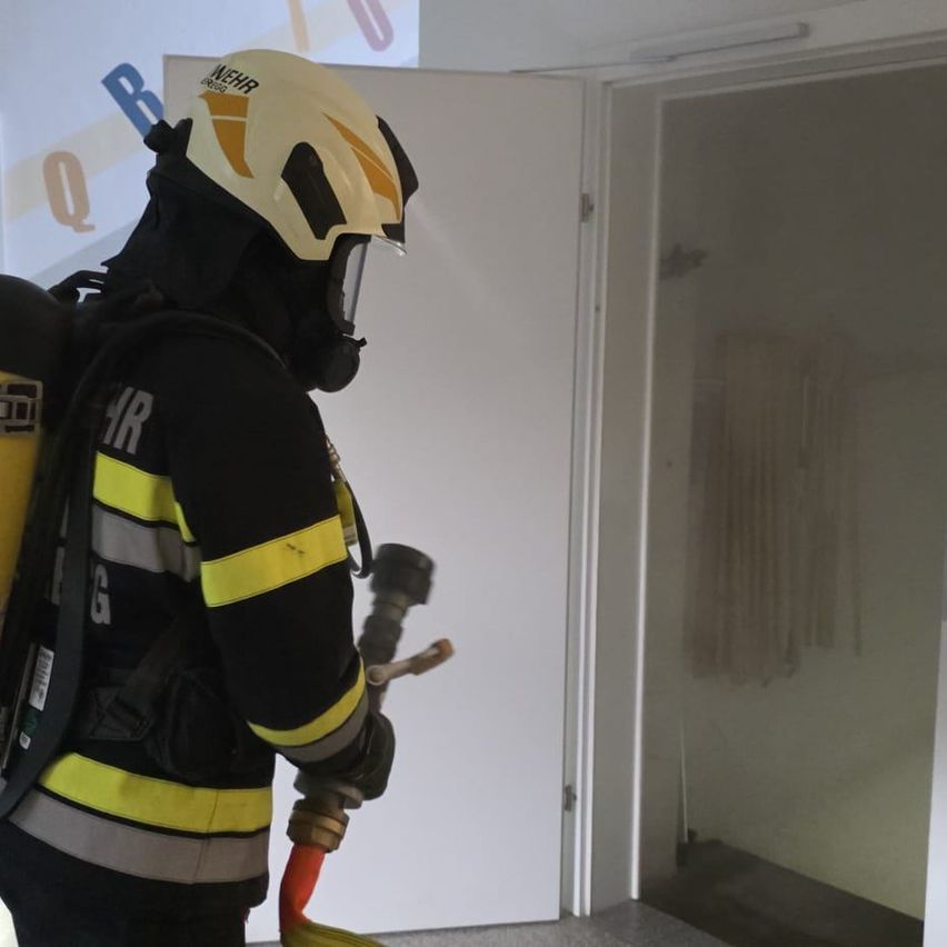 Ein Feuerwehrmann in voller Ausrüstung steht mit einem Feuerlöscher und sprüht Wasser. Eine weiße Tür ist offen, und eine Wand dahinter hat Flecken.