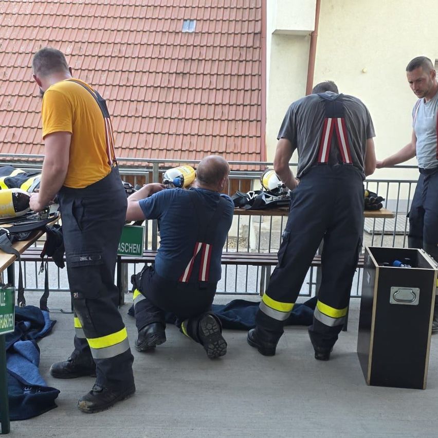 Fünf Feuerwehrleute bereiten ihre Ausrüstung auf einer Bank vor. Einer sitzt mit einem Helm, während ein anderer neben einem Mülleimer steht. Sie tragen Uniformen mit reflektierenden Streifen.