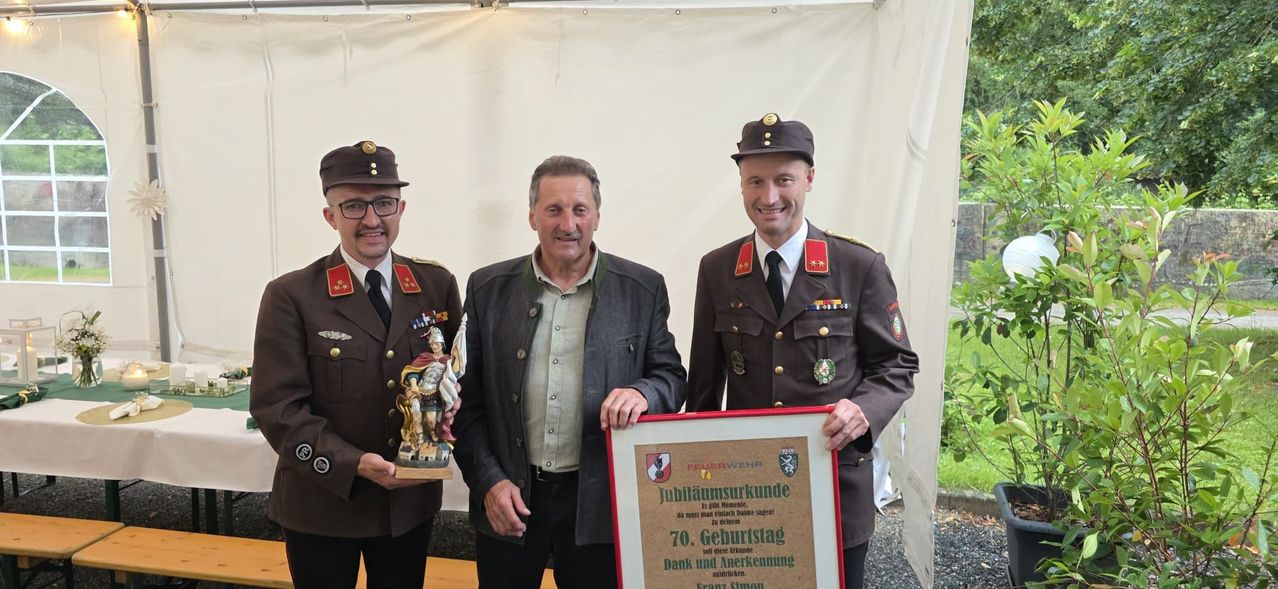 Drei Männer in Uniformen, einer hält ein gerahmtes Zertifikat, der andere eine Figur. Der Mann in der Mitte lächelt, und sie stehen vor einem weißen Hintergrund.