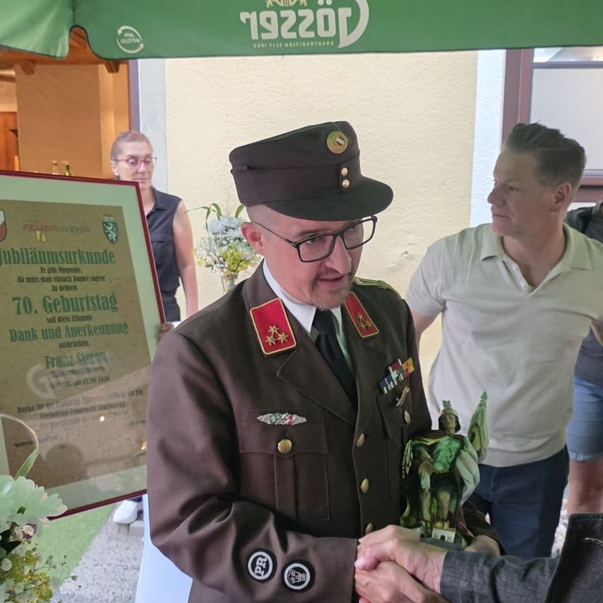 Ein uniformierter Mann hält eine Statue und schüttelt einem anderen Mann die Hand, während eine Frau zusieht, und ein grünes Schild mit Text und Logo im Hintergrund.