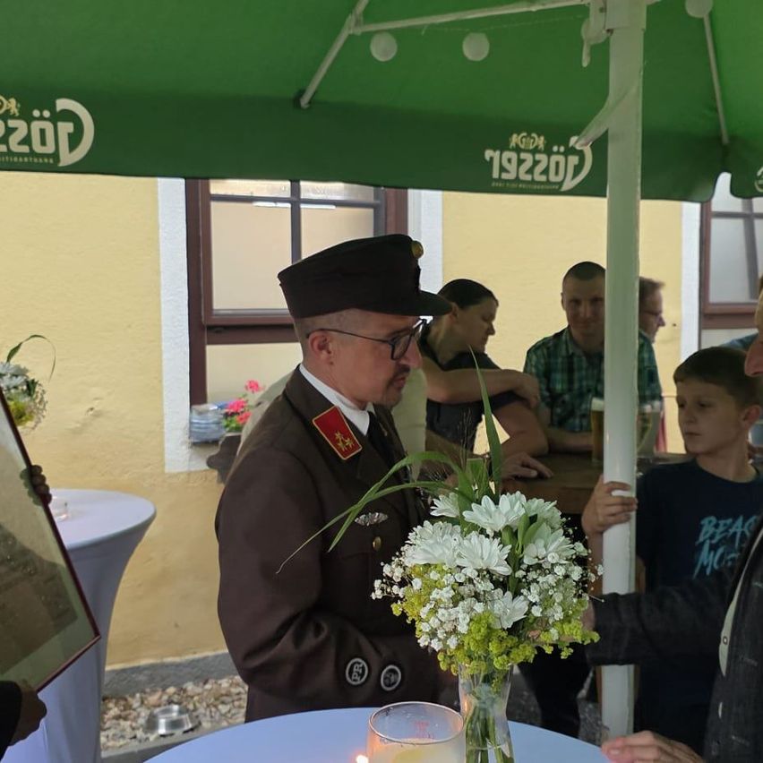 Ein Mann in Uniform steht unter einem grünen Zelt und überreicht einem jungen Jungen eine Blumengestecke. In der Nähe sitzen Menschen an Tischen, wobei eine Person ein Glas Bier hält. Die Szene spielt sich draußen ab.