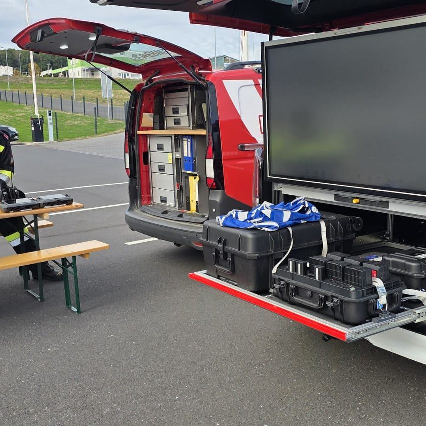 Ein roter Lieferwagen mit offener Heckklappe, der verschiedene Ausrüstungsgegenstände und Aufbewahrungsboxen im Inneren zeigt. Eine Person in Sicherheitsausrüstung sitzt in der Nähe an einem Tisch.