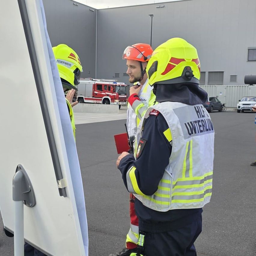 Zwei Feuerwehrleute in gelben Helmen und reflektierenden Westen stehen vor einem Gebäude, einer hält eine rote Mappe. Ein Feuerwehrwagen ist dahinter geparkt.
