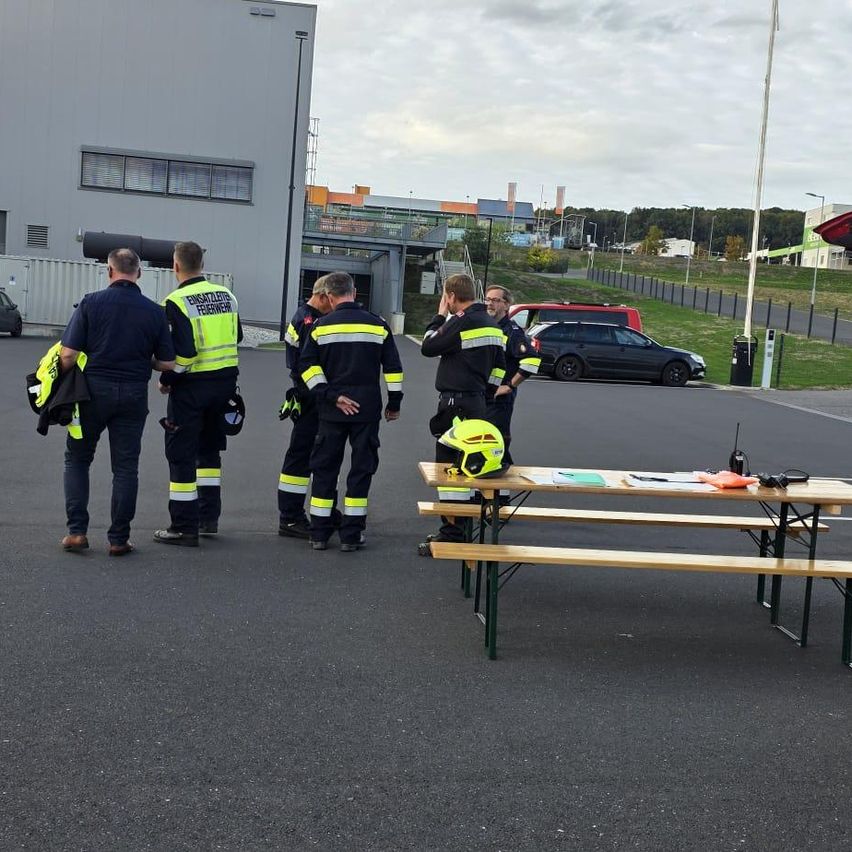 Fünf Feuerwehrleute in reflektierenden Westen stehen auf einem Parkplatz, mit einem Tisch und einem Helm in der Nähe. Ein Gebäude und geparkte Autos sind im Hintergrund.