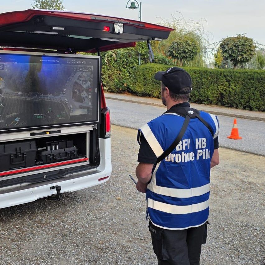 Zwei Feuerwehrleute stehen neben einem roten und weißen Van mit offener Heckklappe. Der Van hat einen großen Monitor auf der Rückseite und ist auf einem Kiesweg geparkt.